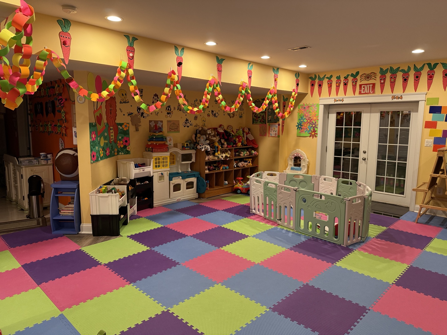 Wide view of the daycare playroom with colorful floor mats, paper chains, toy shelves, and a play enclosure.